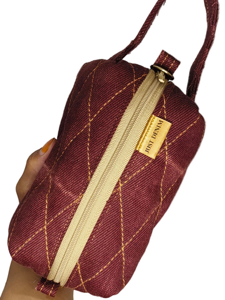 Mini Me Makeup Bag held in one hand, showing burgundy fabric, gold diamond stitching, cream zip and matching loop handle.
